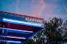 A modern structure with the words 'Electric Corner' prominently displayed in bright lights. The canopy appears metallic with a combination of red and blue accent lighting. The background shows a clear evening sky with some clouds and trees.