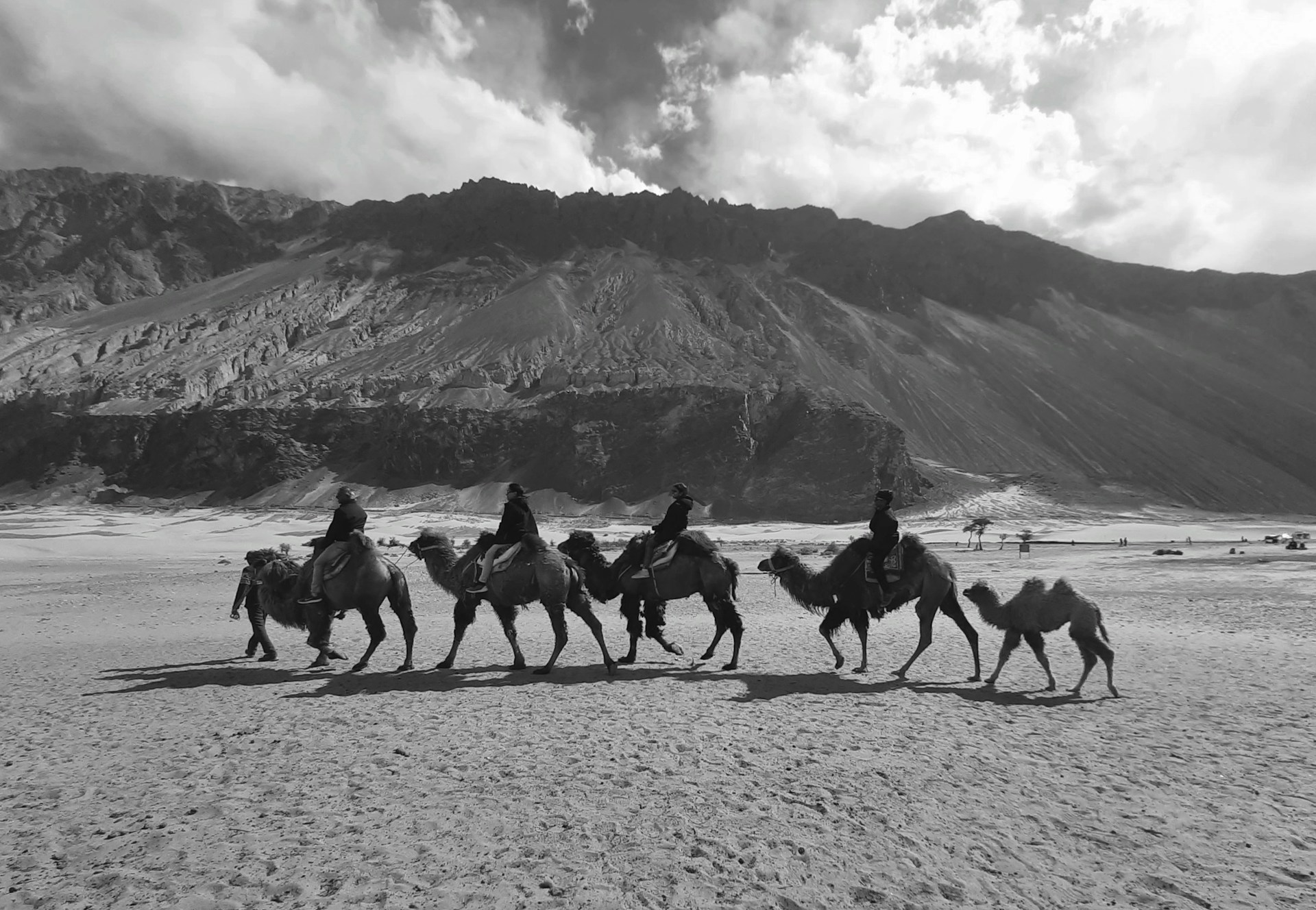 A caravan of camels is being ridden by people across a vast open desert landscape, with a backdrop of towering mountains under a sky filled with scattered clouds.