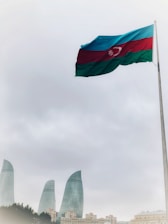 A large flag of Azerbaijan waves prominently on a tall pole set against a cloudy sky. Below, the cityscape features the iconic Flame Towers, strikingly modern skyscrapers made of glass.
