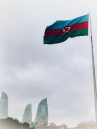 A large flag of Azerbaijan waves prominently on a tall pole set against a cloudy sky. Below, the cityscape features the iconic Flame Towers, strikingly modern skyscrapers made of glass.