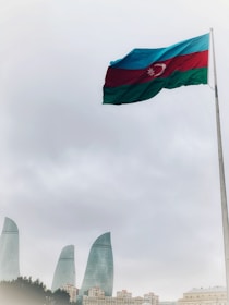 A large flag of Azerbaijan waves prominently on a tall pole set against a cloudy sky. Below, the cityscape features the iconic Flame Towers, strikingly modern skyscrapers made of glass.