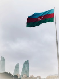 A large flag of Azerbaijan waves prominently on a tall pole set against a cloudy sky. Below, the cityscape features the iconic Flame Towers, strikingly modern skyscrapers made of glass.