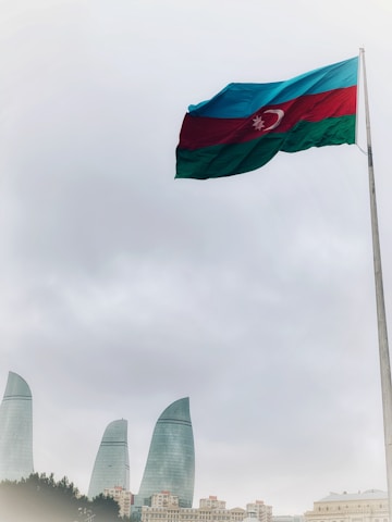 A large flag of Azerbaijan waves prominently on a tall pole set against a cloudy sky. Below, the cityscape features the iconic Flame Towers, strikingly modern skyscrapers made of glass.