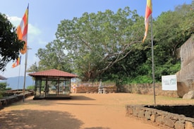 A serene outdoor space featuring a small glass shrine or pavilion with a red-tiled roof. Buddhist flags of various colors are prominently displayed on flagpoles. A large tree with lush, green leaves stands in the background, offering shade to part of the area. A stone fence runs parallel to the tree, with small white structures and vibrant decorations near it. A large rock is situated on the right side.