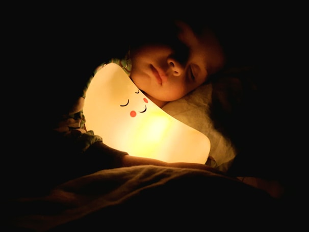 A child peacefully sleeps, holding a glowing night light that resembles a smiling face. The light casts a warm glow, illuminating the child's face and surrounding area in the darkness.