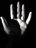 A monochrome image where a human hand is prominently featured, illuminated from one side, casting deep shadows that emphasize the texture and lines of the palm.