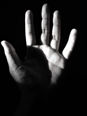 A monochrome image where a human hand is prominently featured, illuminated from one side, casting deep shadows that emphasize the texture and lines of the palm.