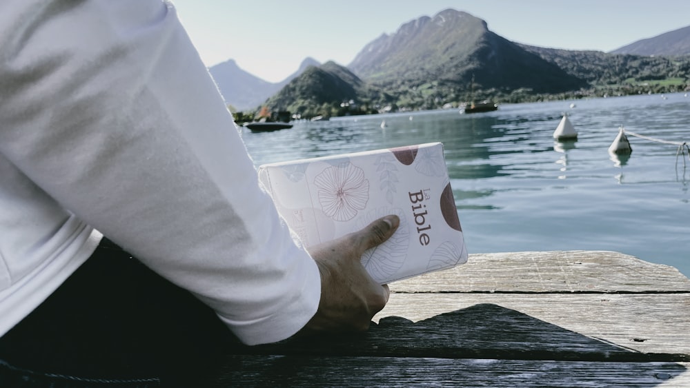 a hand holding a white paper bag over water with mountains in the background