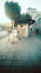 A person wearing a hat and light-colored clothing is sweeping the sidewalk in front of a closed shop with a metal shutter. The scene is set during the day with a tree on the left and a road with moving vehicles on the side. A hotel sign is visible in the background.