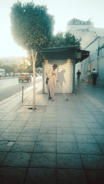 A person wearing a hat and light-colored clothing is sweeping the sidewalk in front of a closed shop with a metal shutter. The scene is set during the day with a tree on the left and a road with moving vehicles on the side. A hotel sign is visible in the background.