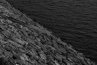 A black and white photograph depicting a rocky shoreline meeting the calm waters of a body of water. The image captures the texture of the rocks and the gentle ripples on the water's surface, with a strong contrast between the two elements.