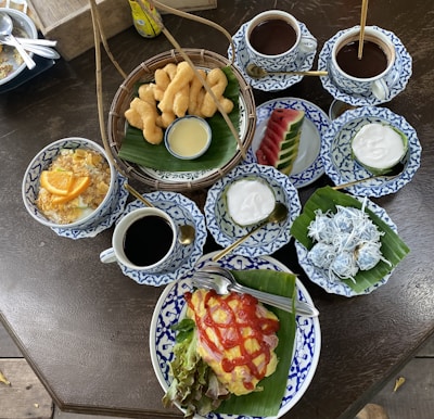 An assortment of beautifully presented dishes on a dark wooden table, featuring an omelette drizzled with ketchup and served with fresh lettuce on a banana leaf. Accompaniments include crispy fried dough sticks with a dipping sauce, fruit slices including watermelon, and small bowls of creamy dessert garnished with shredded coconut. Several cups of coffee or chocolate sit in intricately patterned blue and white ceramic dishes alongside the food.