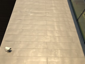 The image features a large expanse of beige-colored tile or concrete surface. There is a security camera mounted on the lower left side. A shadow partially covers the surface on the right, and there are reflective glass windows visible in the top right corner.