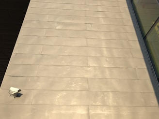 The image features a large expanse of beige-colored tile or concrete surface. There is a security camera mounted on the lower left side. A shadow partially covers the surface on the right, and there are reflective glass windows visible in the top right corner.