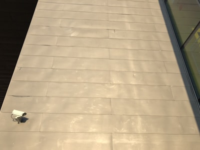 The image features a large expanse of beige-colored tile or concrete surface. There is a security camera mounted on the lower left side. A shadow partially covers the surface on the right, and there are reflective glass windows visible in the top right corner.