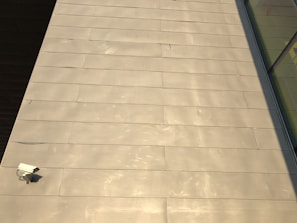 The image features a large expanse of beige-colored tile or concrete surface. There is a security camera mounted on the lower left side. A shadow partially covers the surface on the right, and there are reflective glass windows visible in the top right corner.