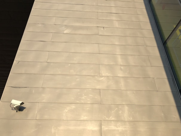 The image features a large expanse of beige-colored tile or concrete surface. There is a security camera mounted on the lower left side. A shadow partially covers the surface on the right, and there are reflective glass windows visible in the top right corner.