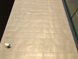 The image features a large expanse of beige-colored tile or concrete surface. There is a security camera mounted on the lower left side. A shadow partially covers the surface on the right, and there are reflective glass windows visible in the top right corner.