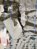 A textured abstract composition featuring various materials layered together, including burlap, paint strokes, and muted colors with hints of blue lines. The surface has a rugged and worn appearance, with smudges and rough patches.