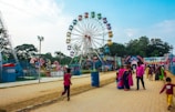 A vibrant outdoor amusement park scene featuring a large Ferris wheel with multicolored gondolas as the central attraction. People of various ages are enjoying the park, with some walking along a paved path and others gathered near rides. The sky is clear, and there are some trees and other amusement rides in the background.