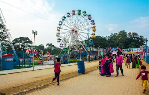 A vibrant outdoor amusement park scene featuring a large Ferris wheel with multicolored gondolas as the central attraction. People of various ages are enjoying the park, with some walking along a paved path and others gathered near rides. The sky is clear, and there are some trees and other amusement rides in the background.