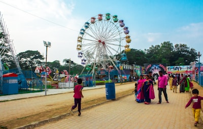 A vibrant outdoor amusement park scene featuring a large Ferris wheel with multicolored gondolas as the central attraction. People of various ages are enjoying the park, with some walking along a paved path and others gathered near rides. The sky is clear, and there are some trees and other amusement rides in the background.