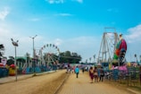 A vibrant amusement park scene with various rides including a large Ferris wheel and a colorful, rocket-shaped ride. The sky is clear and blue, creating a cheerful atmosphere. People are seen walking along a paved pathway, enjoying the attractions and atmosphere.