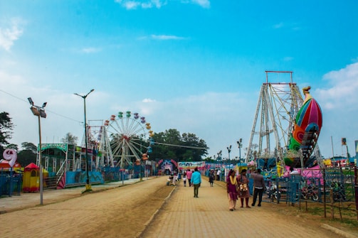 A vibrant amusement park scene with various rides including a large Ferris wheel and a colorful, rocket-shaped ride. The sky is clear and blue, creating a cheerful atmosphere. People are seen walking along a paved pathway, enjoying the attractions and atmosphere.