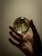 A hand delicately holds a clear glass orb containing a dried flower and chains. The background is softly lit, creating a warm, intimate atmosphere.