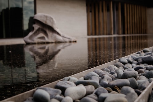 A reflective water surface is in the foreground, with smooth gray pebbles lining the edge. A large abstract stone sculpture is visible across the water, and its reflection creates a symmetrical visual effect. In the background, there are vertical wooden slats and a plain wall, contributing to an overall minimalist design.