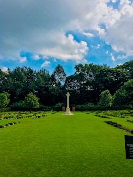 A well-maintained cemetery with a prominent stone cross standing at the center, surrounded by lush greenery and rows of small headstones. The background features dense green trees under a partly cloudy sky with patches of blue.