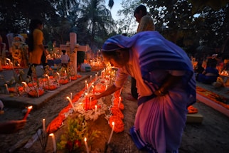 A group of people gather in a cemetery during dusk. The scene is illuminated by numerous candles placed around the graves, some of which are decorated with orange flowers. A woman in traditional attire is seen tending to one of the graves, adjusting the candles or flowers. The background is filled with trees and other silhouettes of people engaged in similar activities.