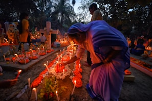A group of people gather in a cemetery during dusk. The scene is illuminated by numerous candles placed around the graves, some of which are decorated with orange flowers. A woman in traditional attire is seen tending to one of the graves, adjusting the candles or flowers. The background is filled with trees and other silhouettes of people engaged in similar activities.