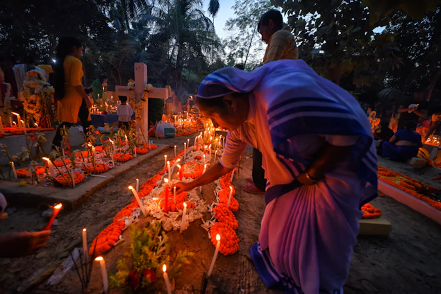 A group of people gather in a cemetery during dusk. The scene is illuminated by numerous candles placed around the graves, some of which are decorated with orange flowers. A woman in traditional attire is seen tending to one of the graves, adjusting the candles or flowers. The background is filled with trees and other silhouettes of people engaged in similar activities.