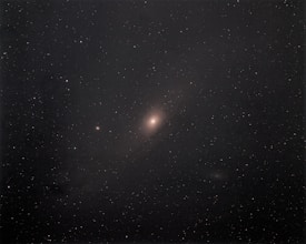A vast array of stars scattered across the night sky, with a prominent glowing elliptical object possibly indicating a galaxy at the center. The surrounding area is dark, highlighting the brightness of the celestial bodies.