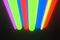 Neon-colored vertical lines in shades of pink, green, red, blue, and yellow on a black background. The colors are vibrant and create a sense of movement.