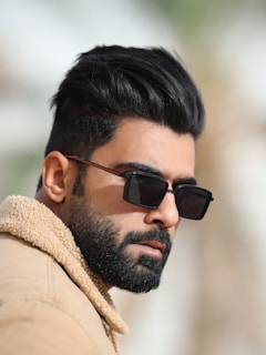 A man with a well-groomed beard and stylish dark hair wears rectangular sunglasses. He is dressed in a tan jacket with a fuzzy collar, looking off to the side in a cool and confident pose.