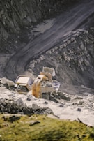 A large excavator is loading rocks into a dump truck on a rugged, rocky quarry terrain. The scene is set against a backdrop of steep, rocky walls and a dirt road, indicating an active mining or construction site.