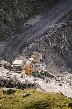 A large excavator is loading rocks into a dump truck on a rugged, rocky quarry terrain. The scene is set against a backdrop of steep, rocky walls and a dirt road, indicating an active mining or construction site.