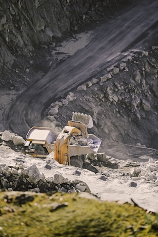 A large excavator is loading rocks into a dump truck on a rugged, rocky quarry terrain. The scene is set against a backdrop of steep, rocky walls and a dirt road, indicating an active mining or construction site.