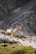 A large excavator is loading rocks into a dump truck on a rugged, rocky quarry terrain. The scene is set against a backdrop of steep, rocky walls and a dirt road, indicating an active mining or construction site.