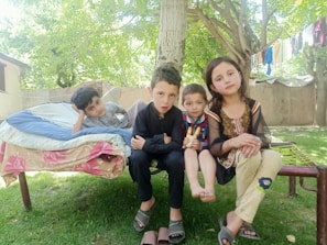 Four children are sitting in an outdoor setting on a traditional wooden charpai, surrounded by lush green trees. One child is lying on his side on a mattress with a floral pattern. The others are sitting side by side, with two looking directly at the camera. Clothes are hanging on a line in the background, and the atmosphere suggests a relaxed, peaceful day.