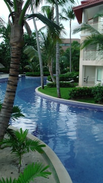 A serene swimming pool curves through a landscaped area surrounded by lush greenery and tall palm trees. Adjacent to the pool is a building with balconies that overlook the scene, bordered by well-maintained grass and small plants. The water in the pool is clear and reflects the green of the trees and blue of the sky.