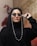 A person with a confident expression wears stylish sunglasses with pearl-adorned straps. The individual is dressed in black attire that includes a headscarf. The backdrop features a beige wall textured with a warm hue. Red leaves are visible in the blurred background, adding contrast.
