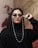 A person with a confident expression wears stylish sunglasses with pearl-adorned straps. The individual is dressed in black attire that includes a headscarf. The backdrop features a beige wall textured with a warm hue. Red leaves are visible in the blurred background, adding contrast.