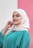 A person wearing a cream-colored hijab and a teal garment is smiling gently while looking at the camera. The background consists of two vertical color blocks in white and pink. The person exhibits a calm and confident demeanor, accessorized with a bracelet.