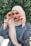 A person wearing a hijab is posed in front of a lush, green foliage background. The individual is dressed in a blue garment and a beige hijab, accessorizing with a watch and bracelets on each wrist. They are looking directly at the camera while adjusting their headscarf on either side.