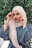 A person wearing a hijab is posed in front of a lush, green foliage background. The individual is dressed in a blue garment and a beige hijab, accessorizing with a watch and bracelets on each wrist. They are looking directly at the camera while adjusting their headscarf on either side.