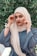 A person wearing a hijab is posed in front of a lush, green foliage background. The individual is dressed in a blue garment and a beige hijab, accessorizing with a watch and bracelets on each wrist. They are looking directly at the camera while adjusting their headscarf on either side.