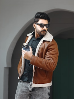 A man with dark hair and a beard is wearing sunglasses, a brown shearling jacket, and a black shirt. He is holding a bottle of cologne in his right hand. He stands in front of an archway, and his posture is slightly angled to the side.